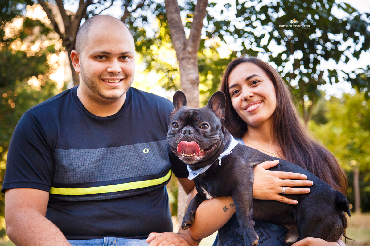 Foto de um cão da raça bulldog francês com seus tutores. Todos olham para a foto sorrindo, o cão com a língua para fora.