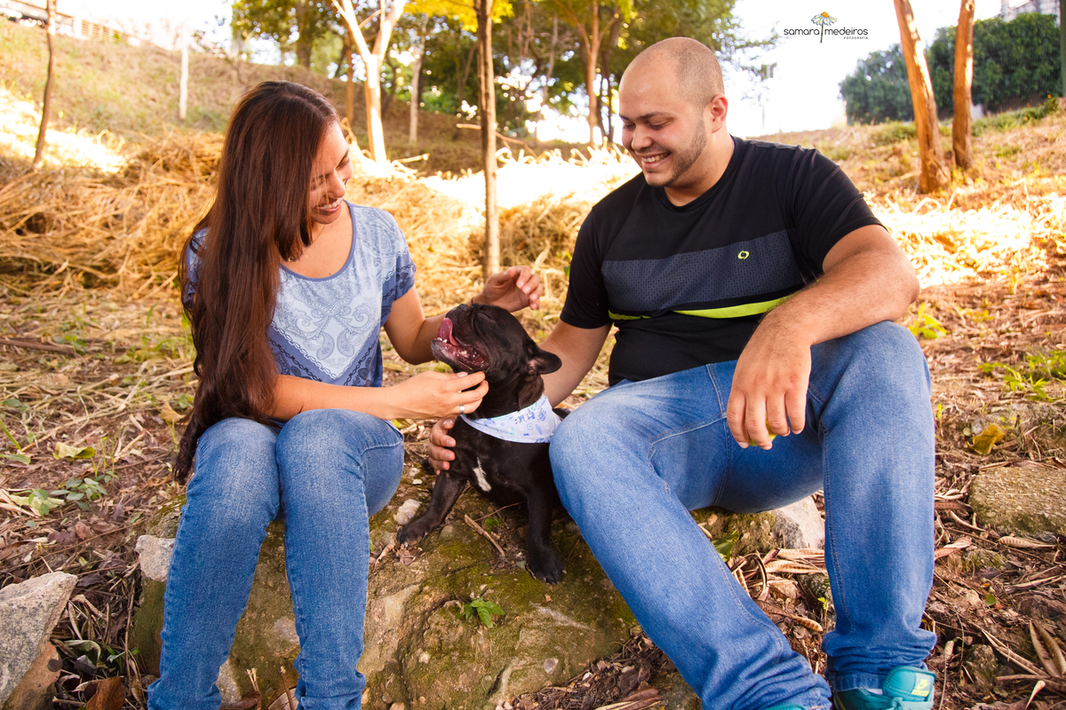 Casal sentado em uma pedra, em um parque de Belo Horizonte, com seu cão da raça bulldog francês entre eles, ambos o fazem carinho, olhando para ele e sorrindo.
