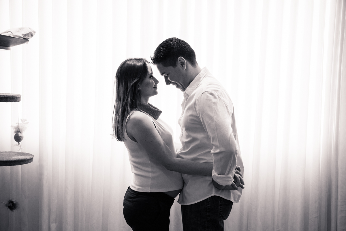 Foto em preto e branco de um casal, no qual a mulher está grávida, se abraçando, de frente um para o outro e se olhando.