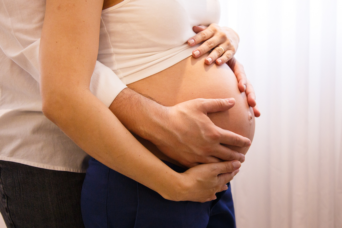 Close na barriga da gestante com suas duas mãos acariciando a barriga e as duas mãos do marido também.