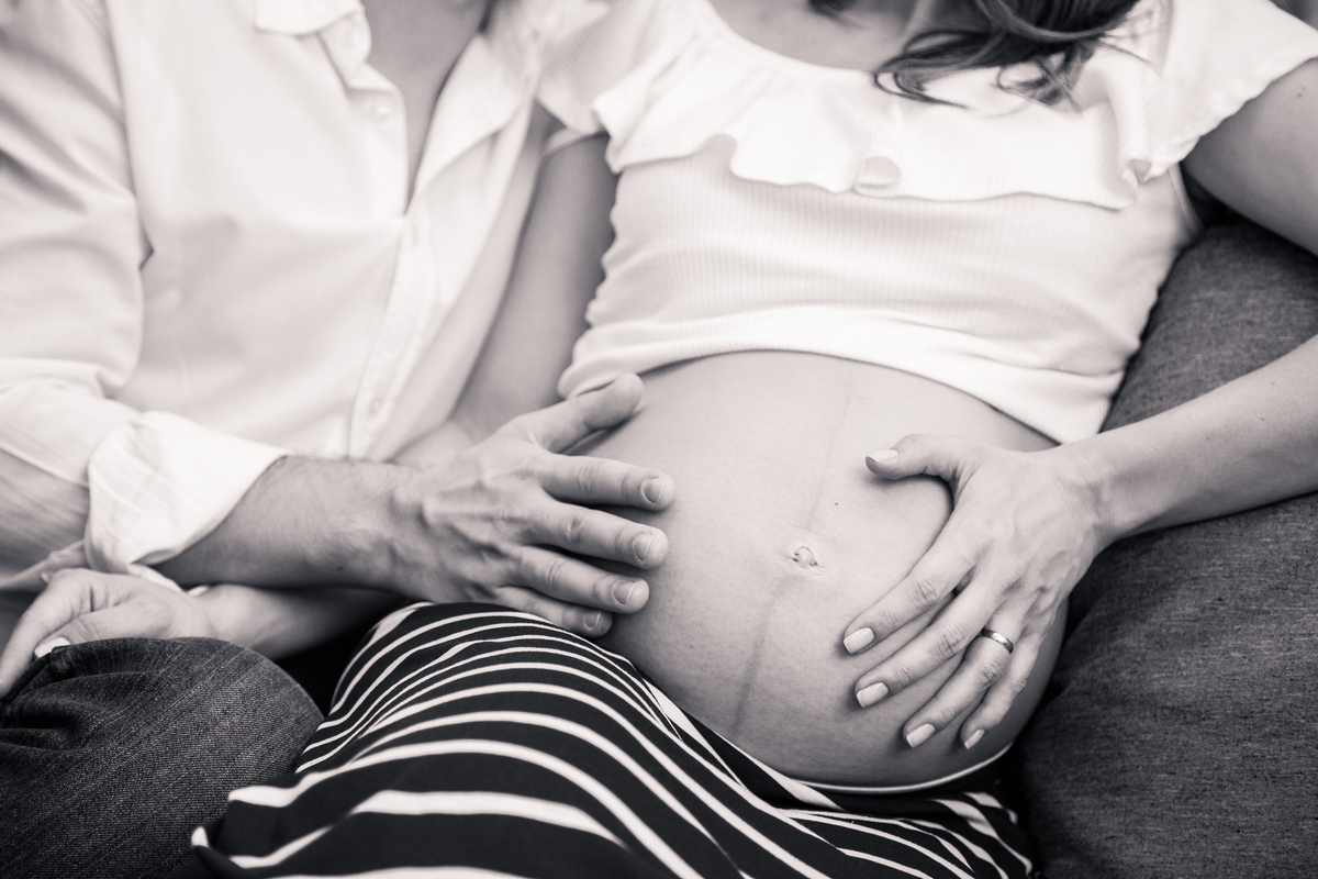 Foto em preto e branco de uma gestante e seu marido sentados no sofá, a foto pega somente do ombro para baixo, uma das mãos da gestante e uma das mãos do marido estão na barriga dela.