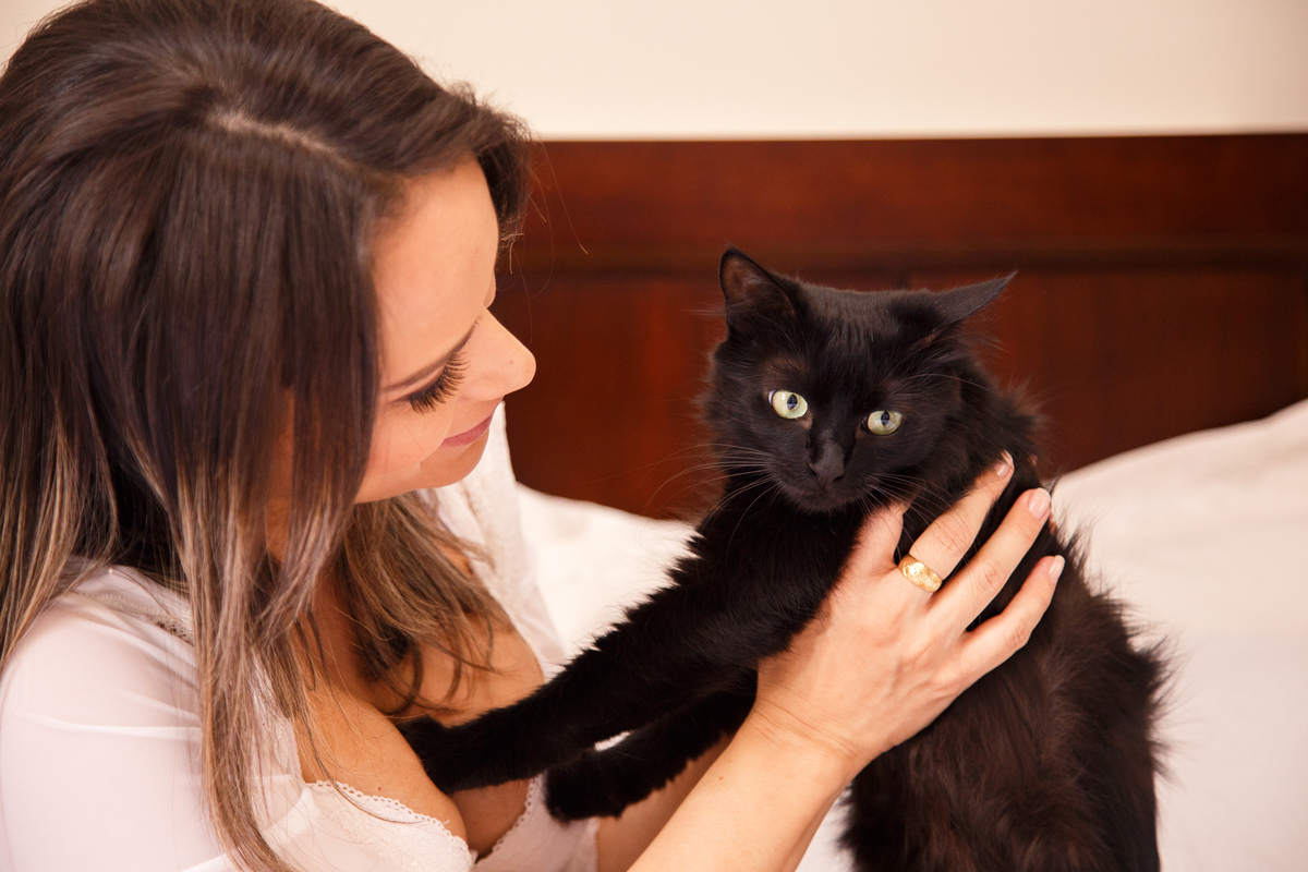 Mulher gestante sentada na cama com sua gatinha nos braços. A gestante olha para a gatinha que olha na direção da câmera.