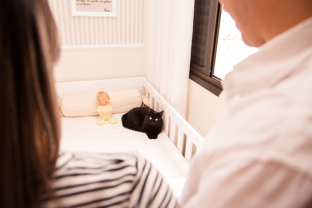 Ao fundo, focada, uma gatinha preta de olhos verdes deitada no berço da irmã humana que está para chegar. Em primeiro plano, desfocados, os tutores de costas, olhando para a gatinha.