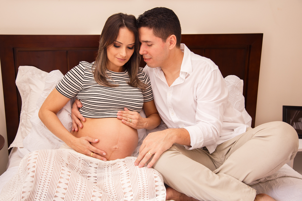 Casal, a mulher grávida, sentados em sua cama, com as pernas cruzadas. A gestante acaricia a barriga e o marido a abraça pelas costas e apoia o rosto no seu.
