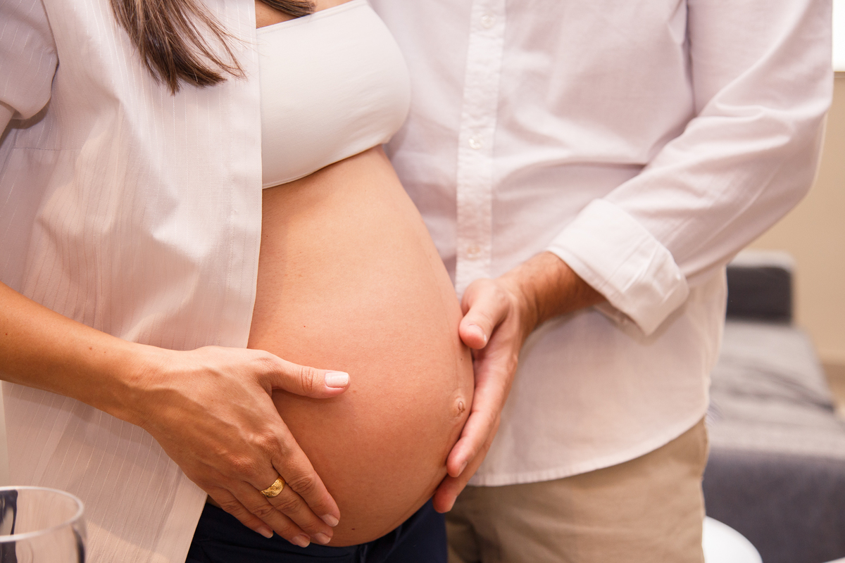 Foto de um casal gestante, com um corte do ombro para baixo, aparecendo o tronco e barriga da gestante. Cada um apoia uma mão na barriga, fazendo carinho.
