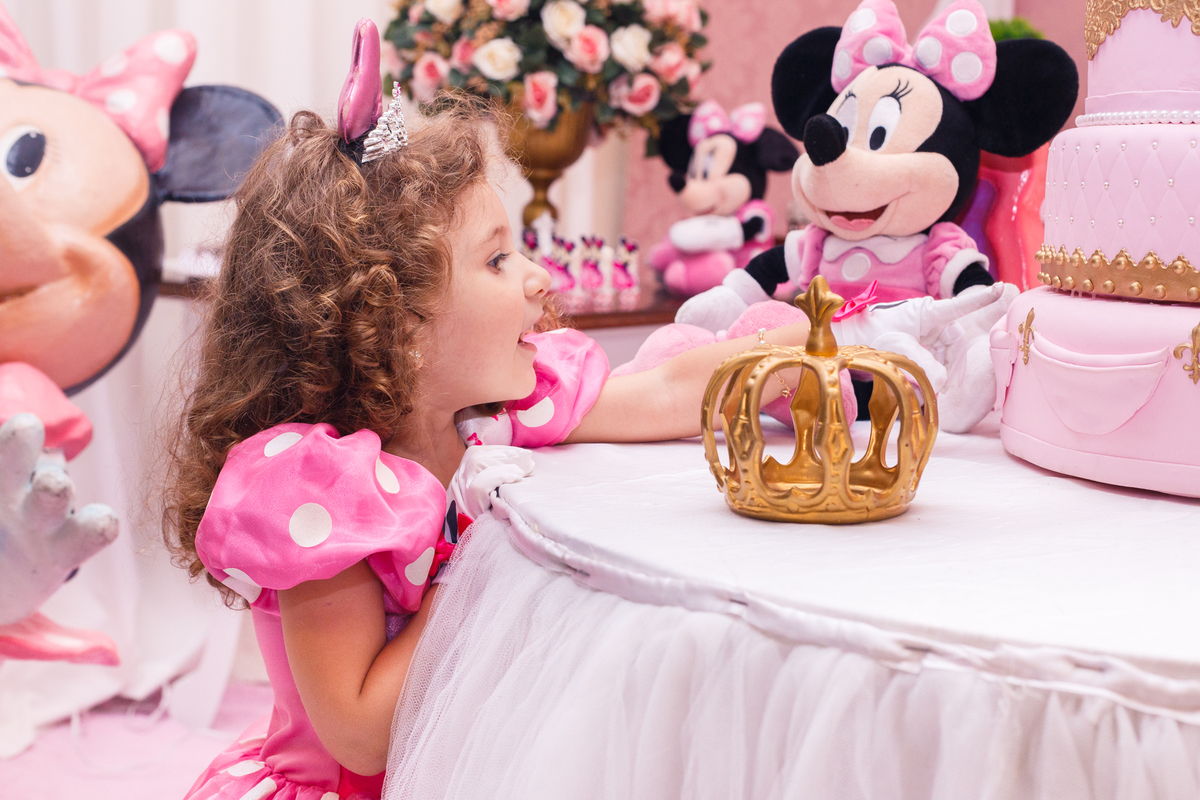 Criança em seu aniversário de 3 anos olhando por cima da mesa dos doces com o braço esticado tentando encostar o dedo no bolo cenográfico.