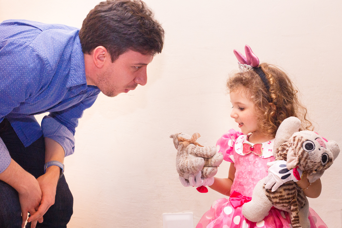 Criança com algumas pelúcias no colo mostrando uma delas para o tio que está agachado conversando com ela.