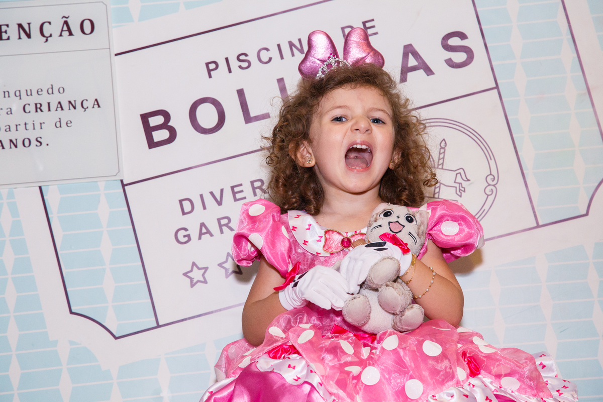 Criança sentada na plataforma que derruba dentro da piscina de bolinhas, com a boca bem aberta fazendo gracinhas e abraçada com um urso de pelúcia.