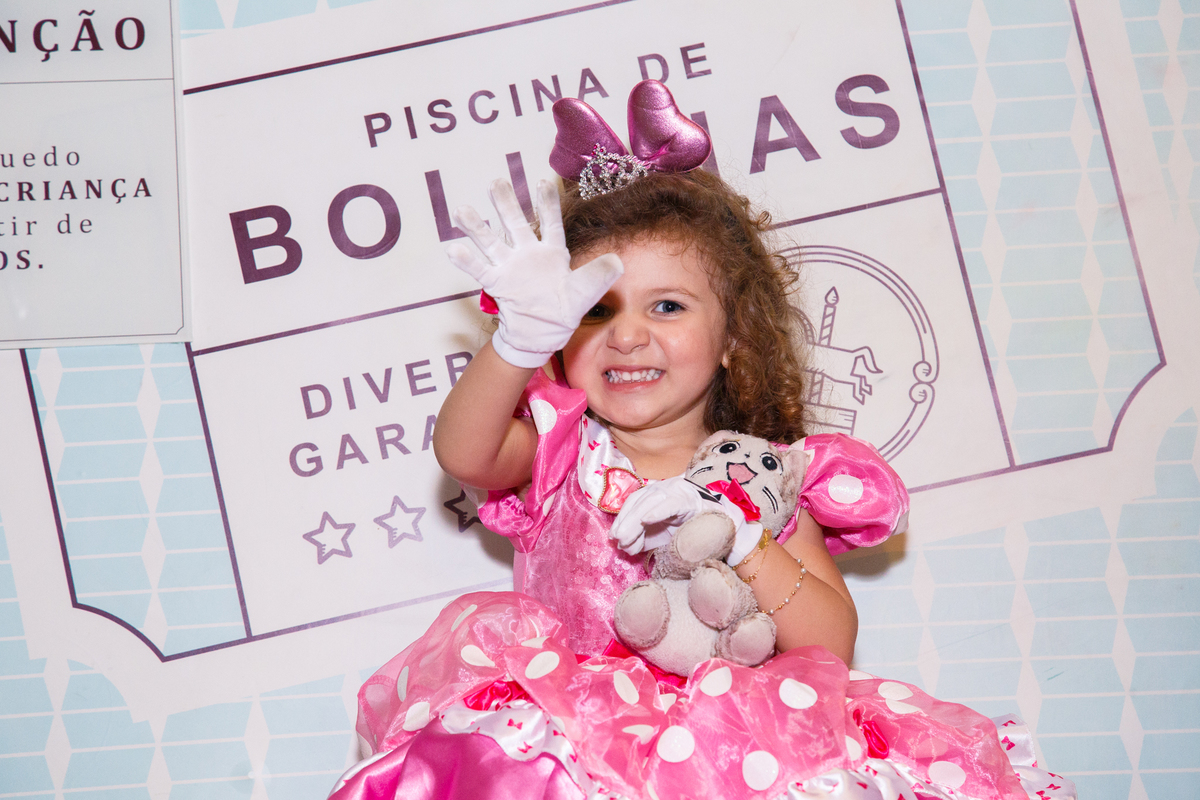 Criança sentada na plataforma que derruba dentro da piscina de bolinhas, sorrindo e dando um tchauzinho, abraçada com um bichinho de pelúcia.