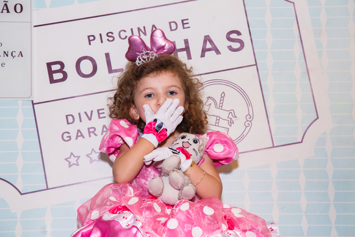 Criança sentada na plataforma que derruba dentro da piscina de bolinhas, mandando um beijo com a mão e abraçada com um bichinho de pelúcia.