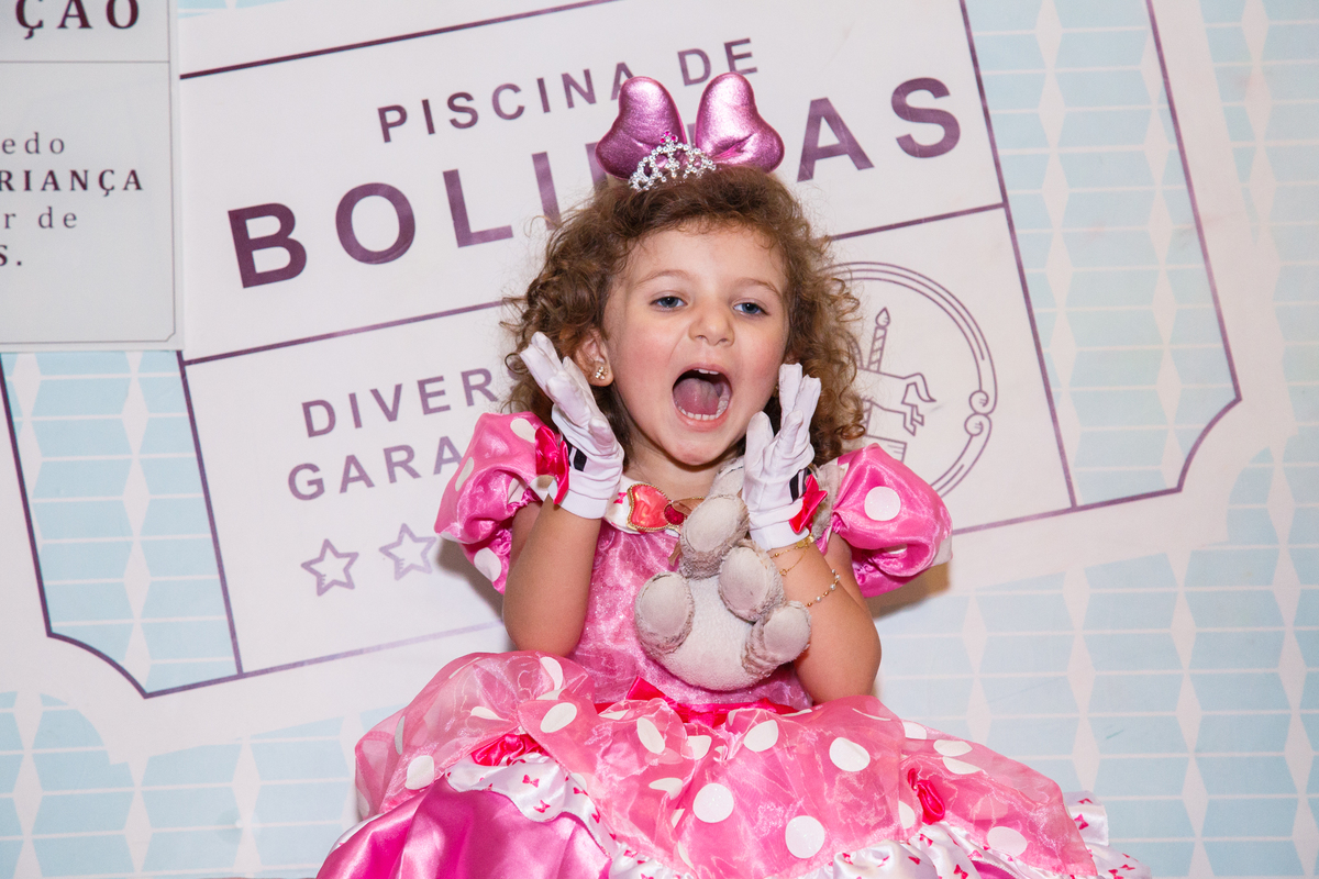 Criança sentada na plataforma que derruba dentro da piscina de bolinhas, com a boca bem aberta, mandando um beijo com as duas mãos e segurando um bichinho de pelúcia.