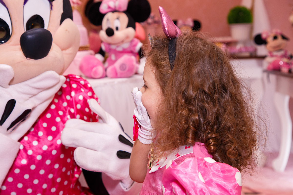 Criança em seu aniversário infantil brincando com a personagem animada Minnie, as duas imitando uma à outra com a mão na boca.