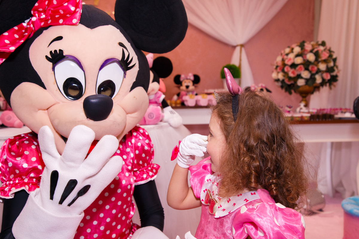 Foto da criança em seu aniversário infantil junto com a personagem animada Minnie, as duas fazendo caretas e brincando, a Minnie com a mão na boca e a criança segurando o próprio nariz e rindo.