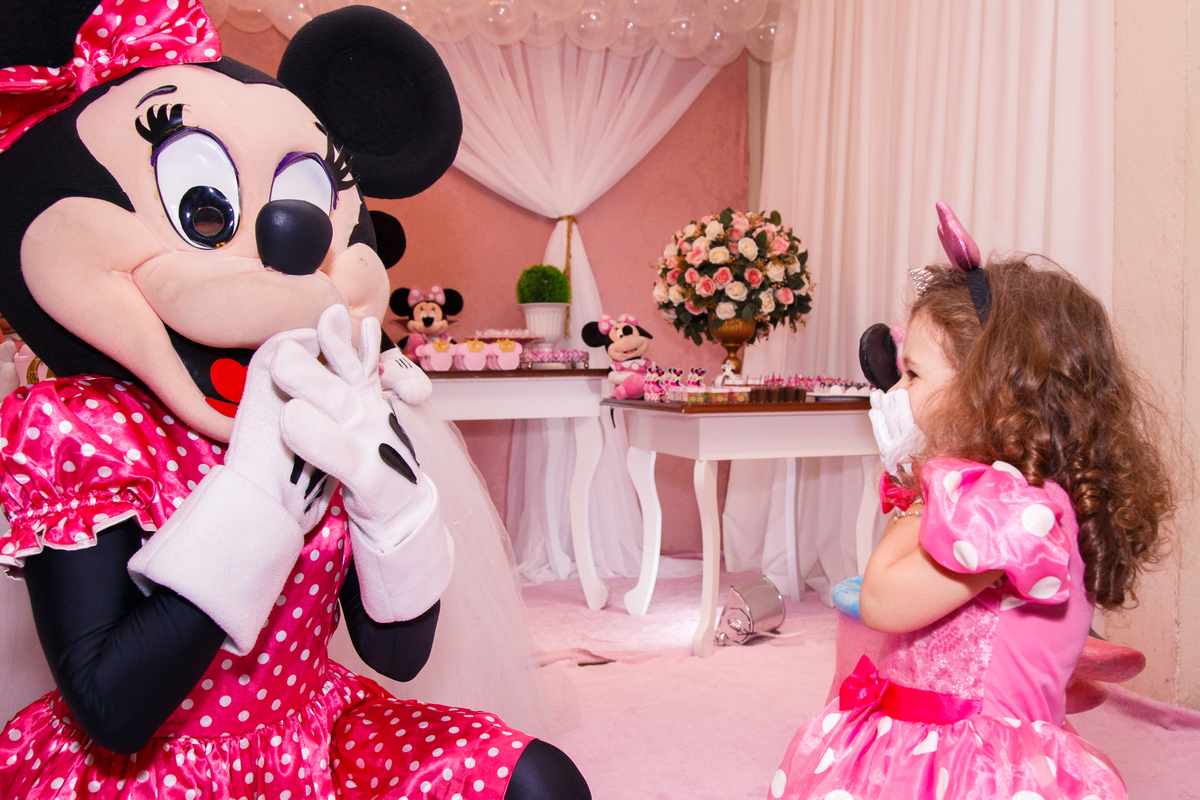 Foto da criança em seu aniversário infantil junto com a personagem animada Minnie as duas imitando uma à outra, com as duas mãos cobrindo a boca.