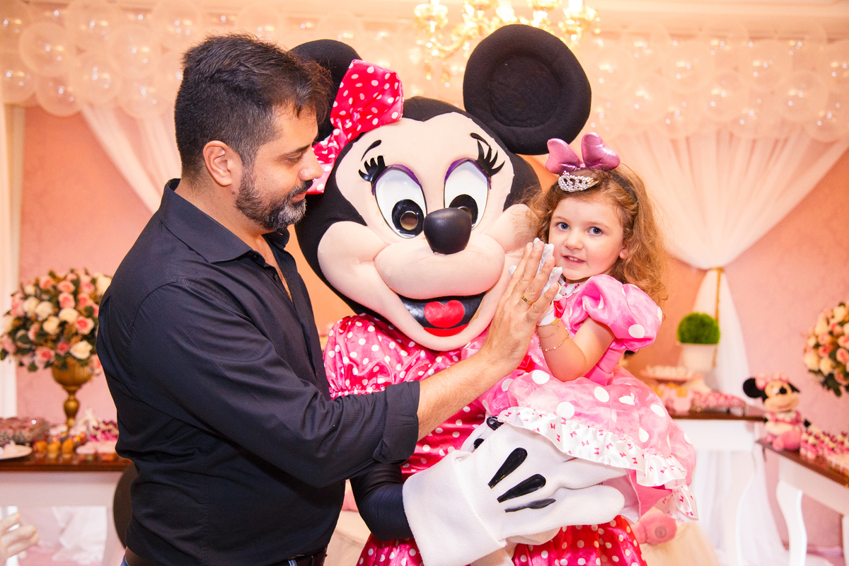 Criança e seu pai junto com a personagem animada Minnie em seu aniversário infantil em um buffet de Belo Horizonte. A criança e seu pai estão com as mãos se tocando, ela no colo da Minnie.