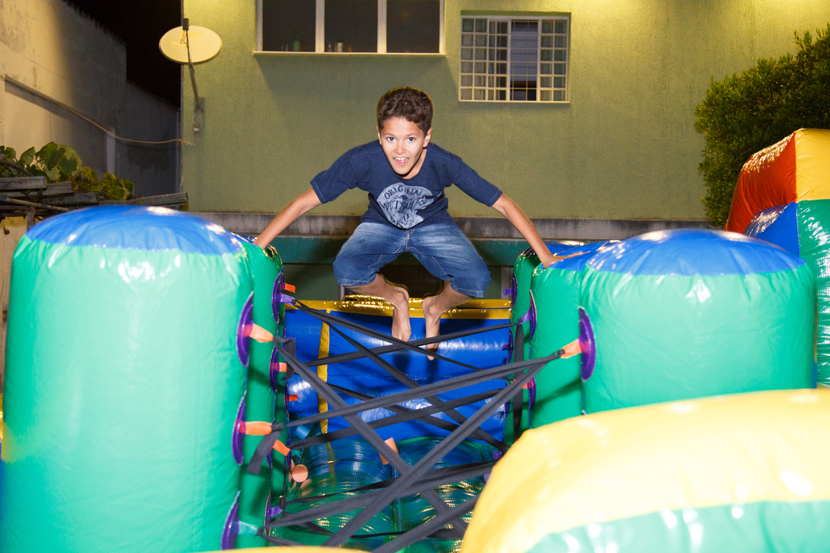Criança brincando no brinquedão inflável na parte de obstáculos dando um salto para conseguir ultrapassar a barreira de cordas, olhando para a câmera e fazendo uma careta.
