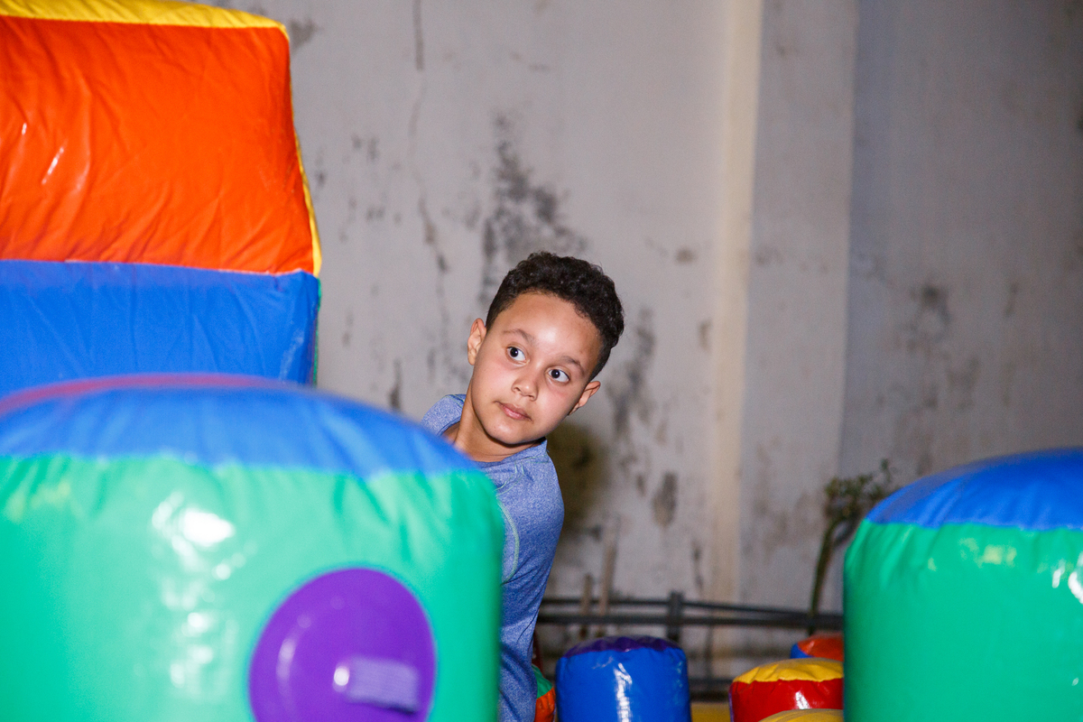 Criança na fila para a rampa do escorrega do brinquedão inflável, ele olha para o lado, parcialmente escondido pela estrutura do brinquedo.