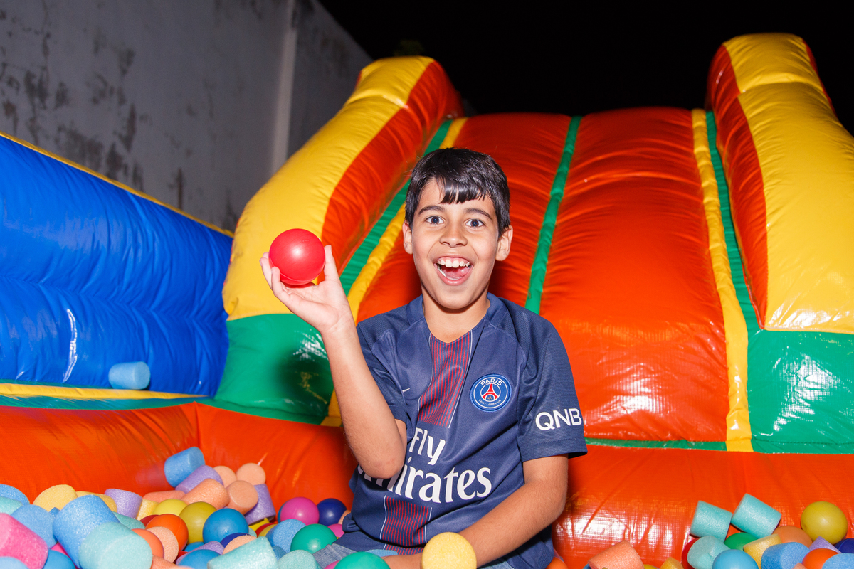 Criança em seu aniversário de 9 anos dentro da piscina de bolinhas do brinquedão inflável com uma bolinha na mão fazendo que vai jogar na direção da câmera, com um sorriso sapeca.