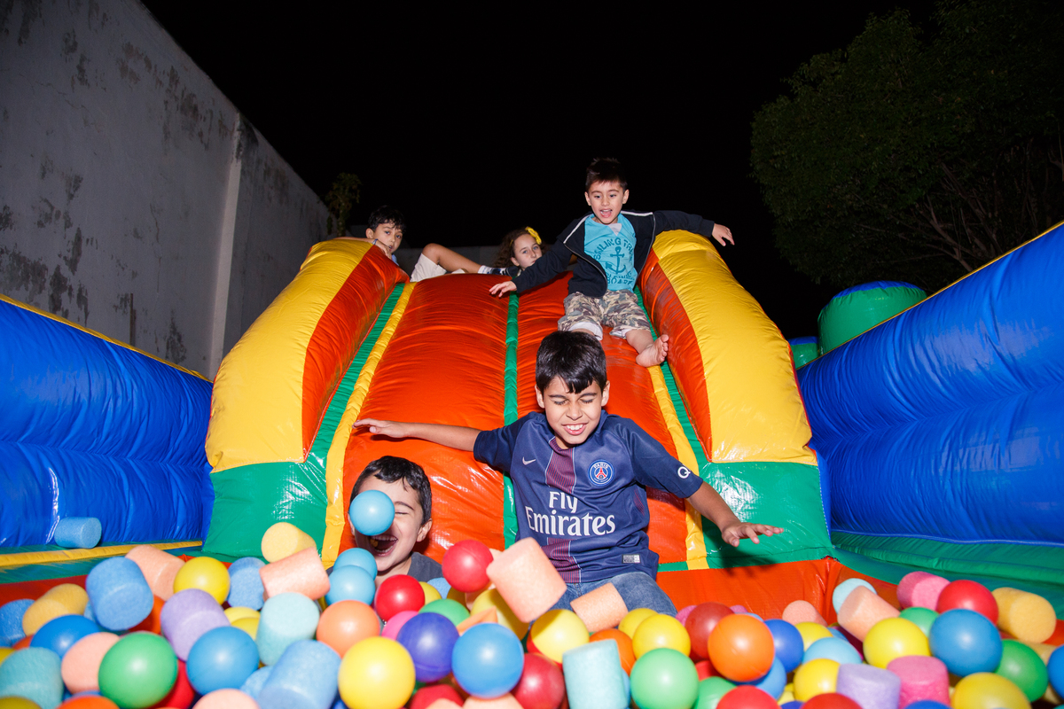 Três crianças escorregando na rampa para a piscina de bolinhas do brinquedão inflável em aniversário infantil.