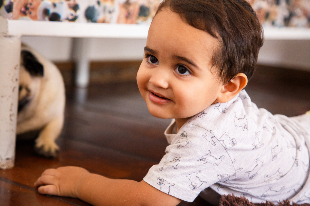 Menino deitado no chão de sua casa brincando com o cão da família da raça pug, que não aparece por completo na foto.