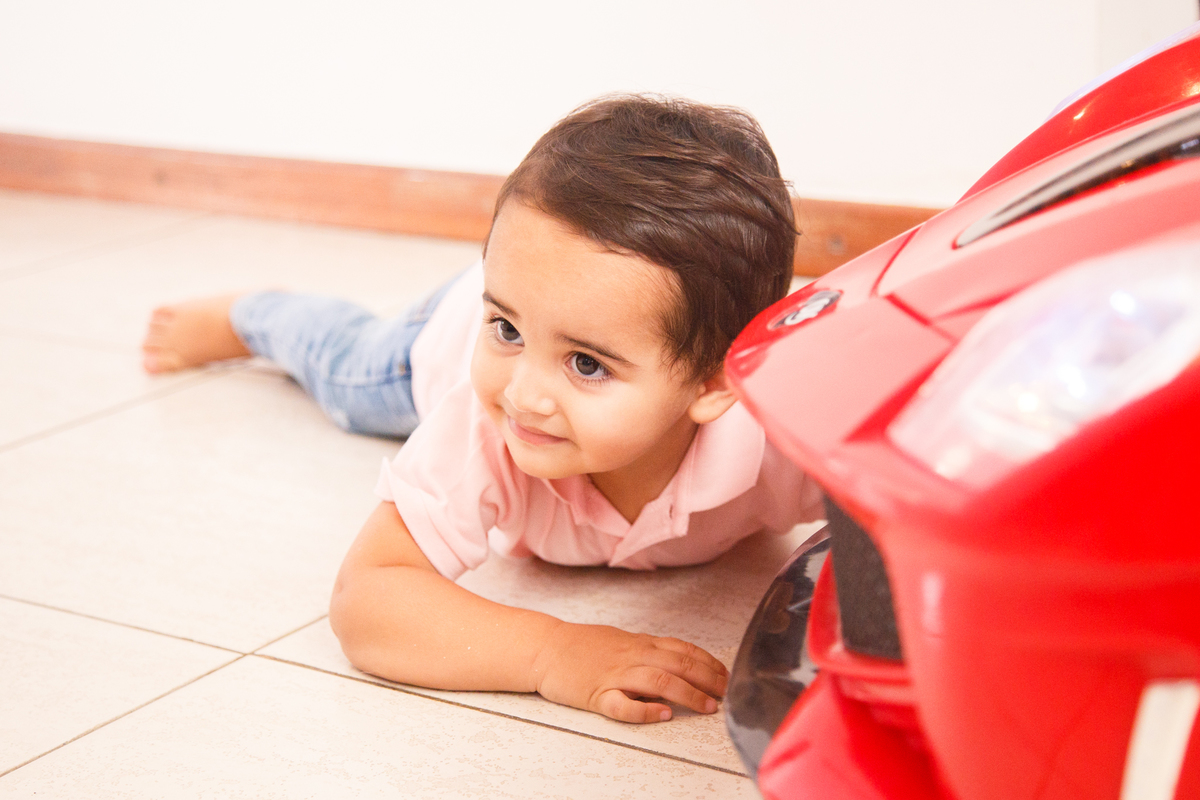 Menino deitado no chão ao lado de seu carro de brinquedo, que aparece parcialmente na foto, em primeiro plano, com a criança em foco no segundo plano.