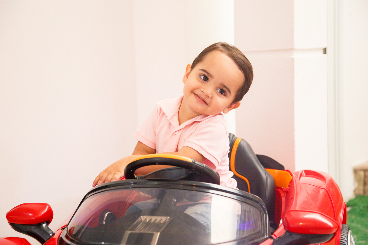 Criança dentro do seu carrinho de brinquedo vermelho, segurando o volante e girando, com o corpo levemente tombado para o lado e olhando de lado para sua mãe que não aparece na foto.