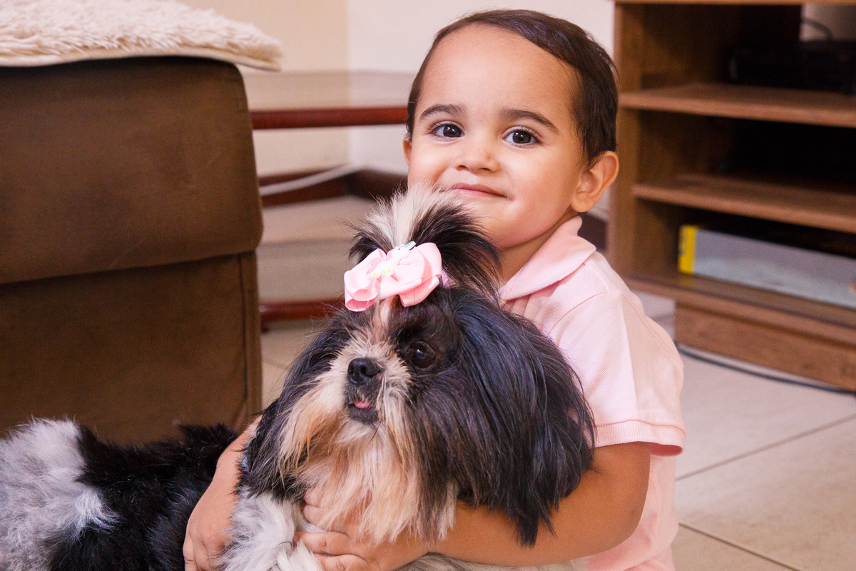 Menino sentado no chão de sua casa abraçado com um dos cães da família, uma fêmea preta e branca da raça shih tzu.
