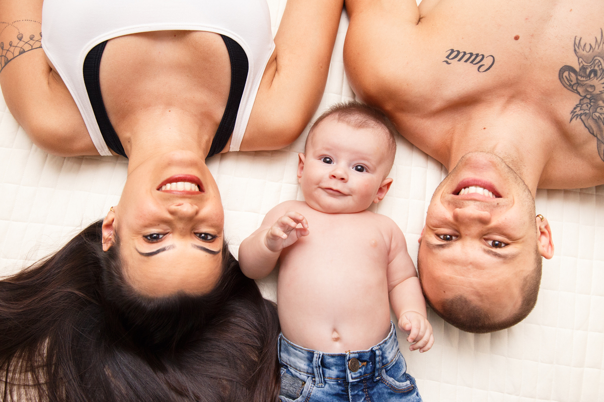 Casal deitado de barriga para cima, olhando para a câmera e sorrindo com seu bebê também deitado olhando para cima entre eles.