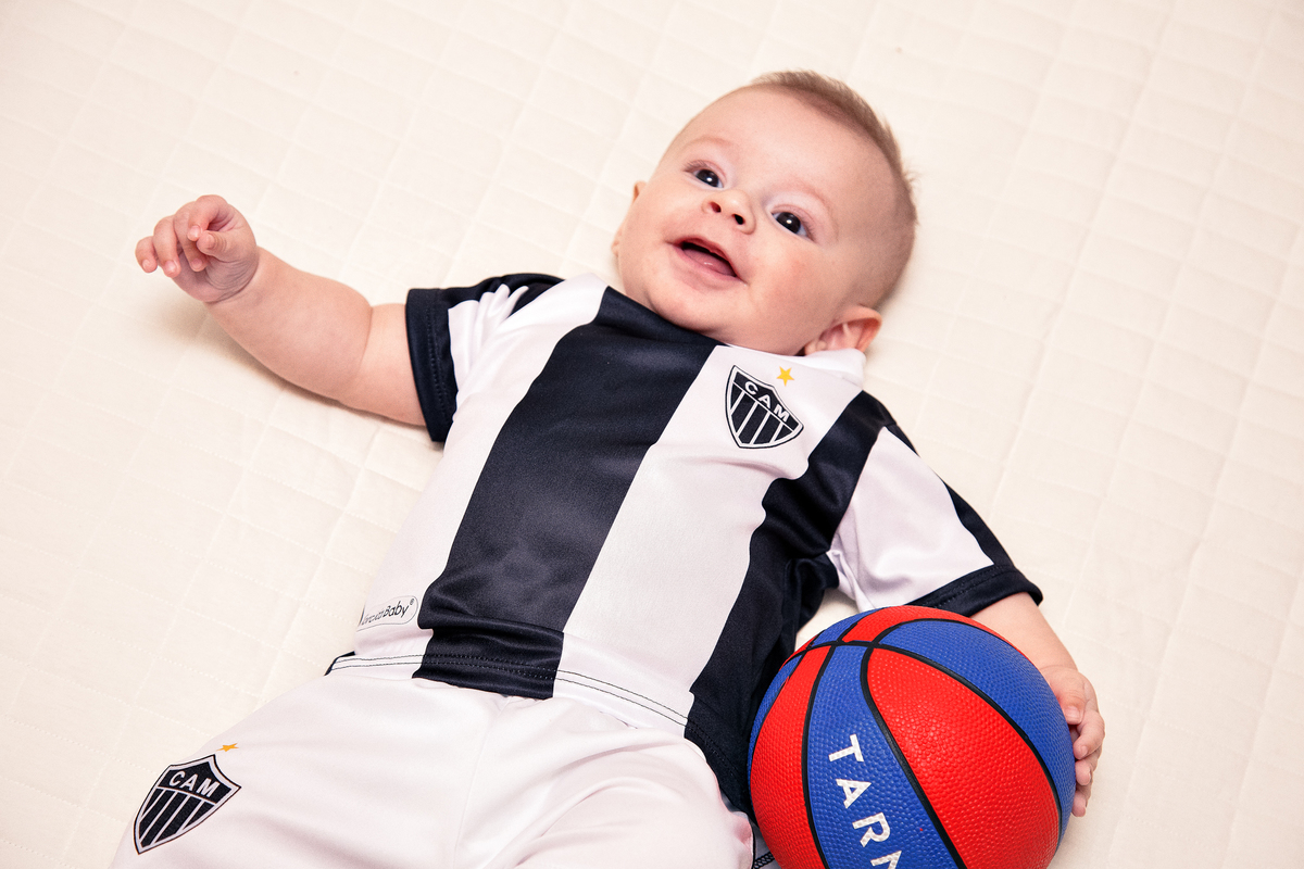 Bebê deitado na cama de barriga para cima sorrindo para os pais e segurando uma bola.