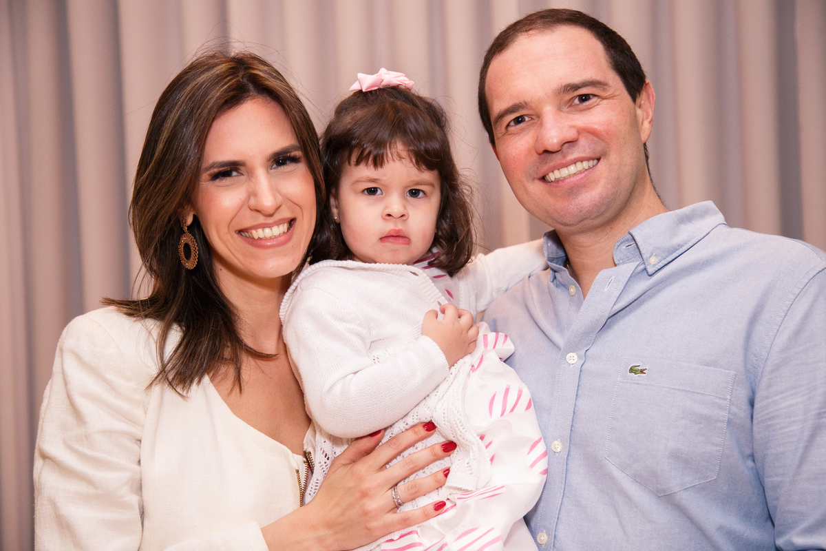 Mãe e pai com a filha de 2 anos no colo, todos olhando para a camera. Pai e mãe sorrindo e a menina séria.