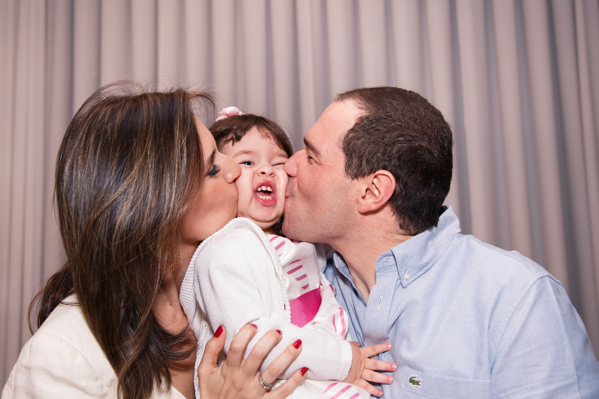 Mãe e pai com a filha de 2 anos no colo, a filha no meio e os dois dando um beijo nela, um de cada lado. Ela faz uma careta.