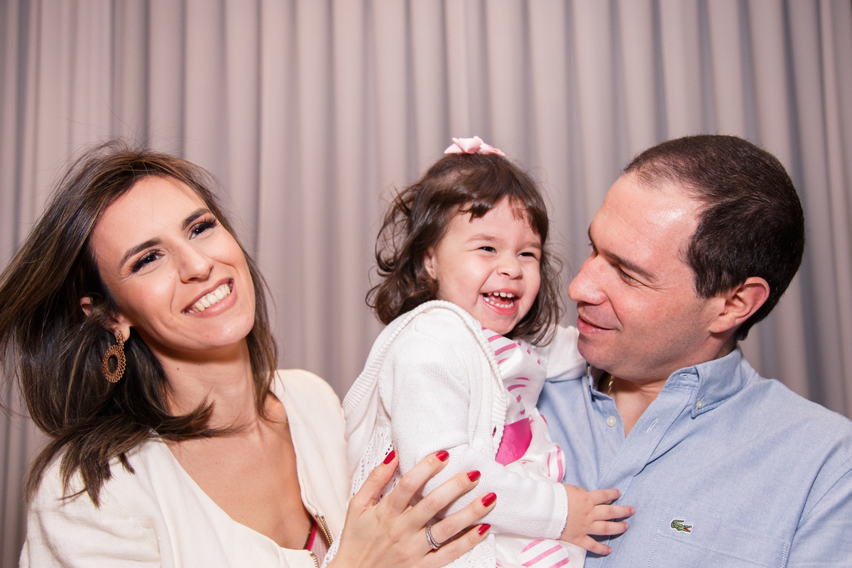 Mãe e pai com a filha de 2 anos no colo, todos sorrindo, o pai olha para  a filha que está entre os dois.