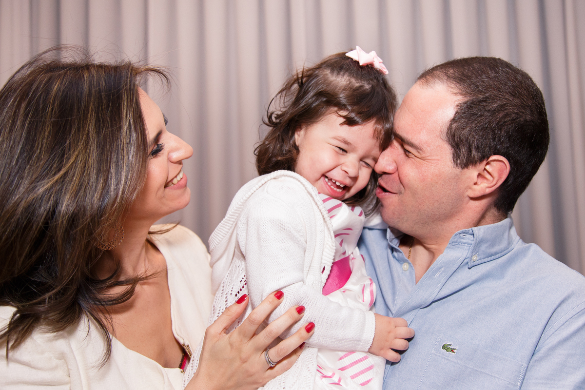 Mãe e pai com a filha de 2 anos no colo, todos sorrindo, o pai está com o rosto encostado no da filha e a mãe olha para os dois.