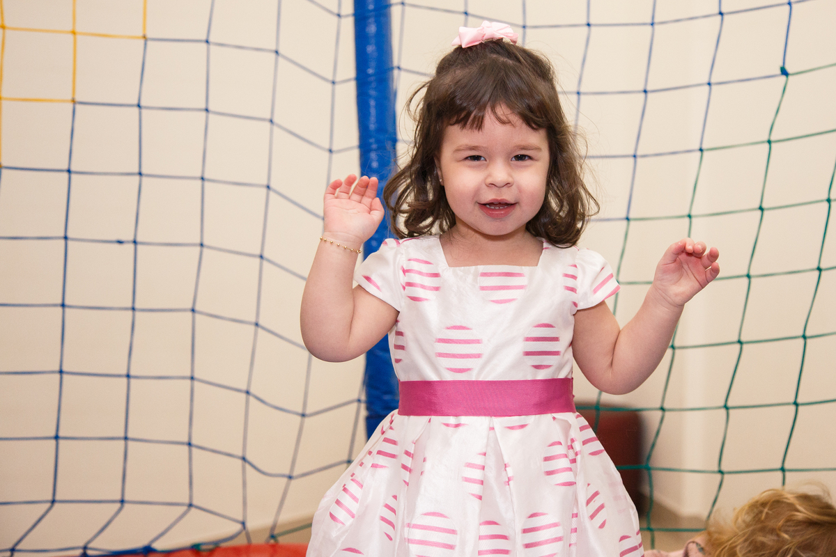 Menina de 2 anos pulando no pula pula em sua festa de aniversário com os dois braços levantados e sorrindo com os olhos meio fechados. Usa um vestido branco com detalhes rosa.