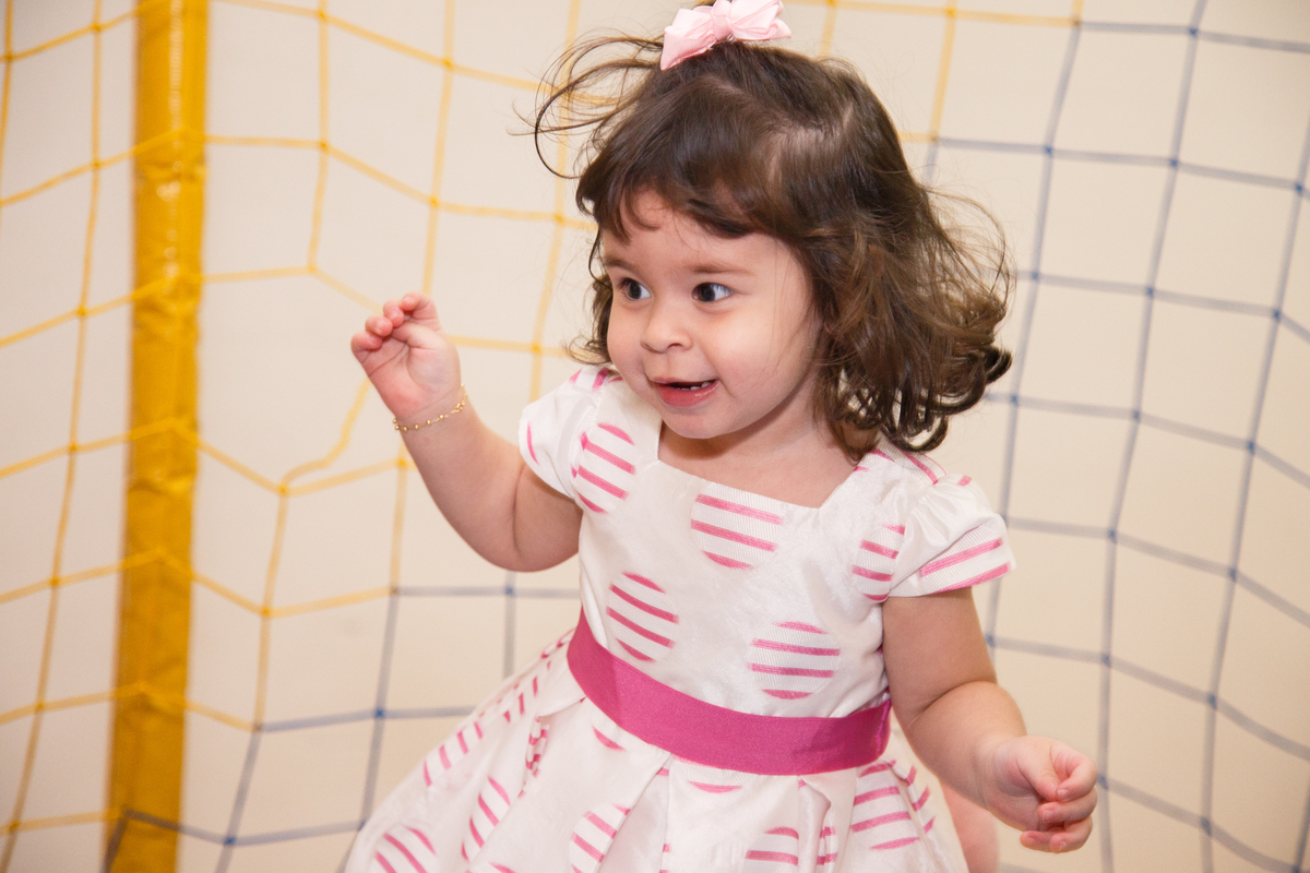 Menina de 2 anos pulando no pula pula em sua festa de aniversário com os dois braços levantados e sorrindo e olhando para o lado. Usa um vestido branco com detalhes rosa.