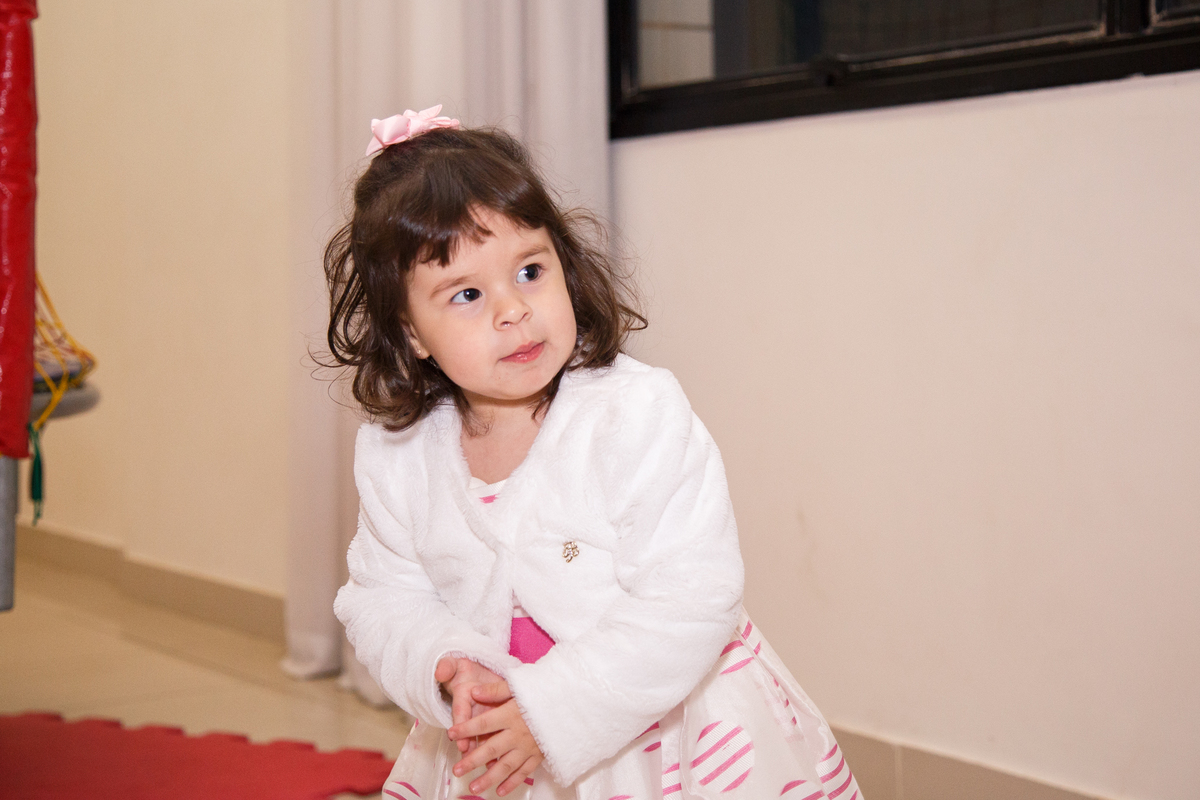 Menina de 2 anos dançando e batendo palma no meio da foto, usando um vestido branco com detalhes rosa e um casaquinho branco.