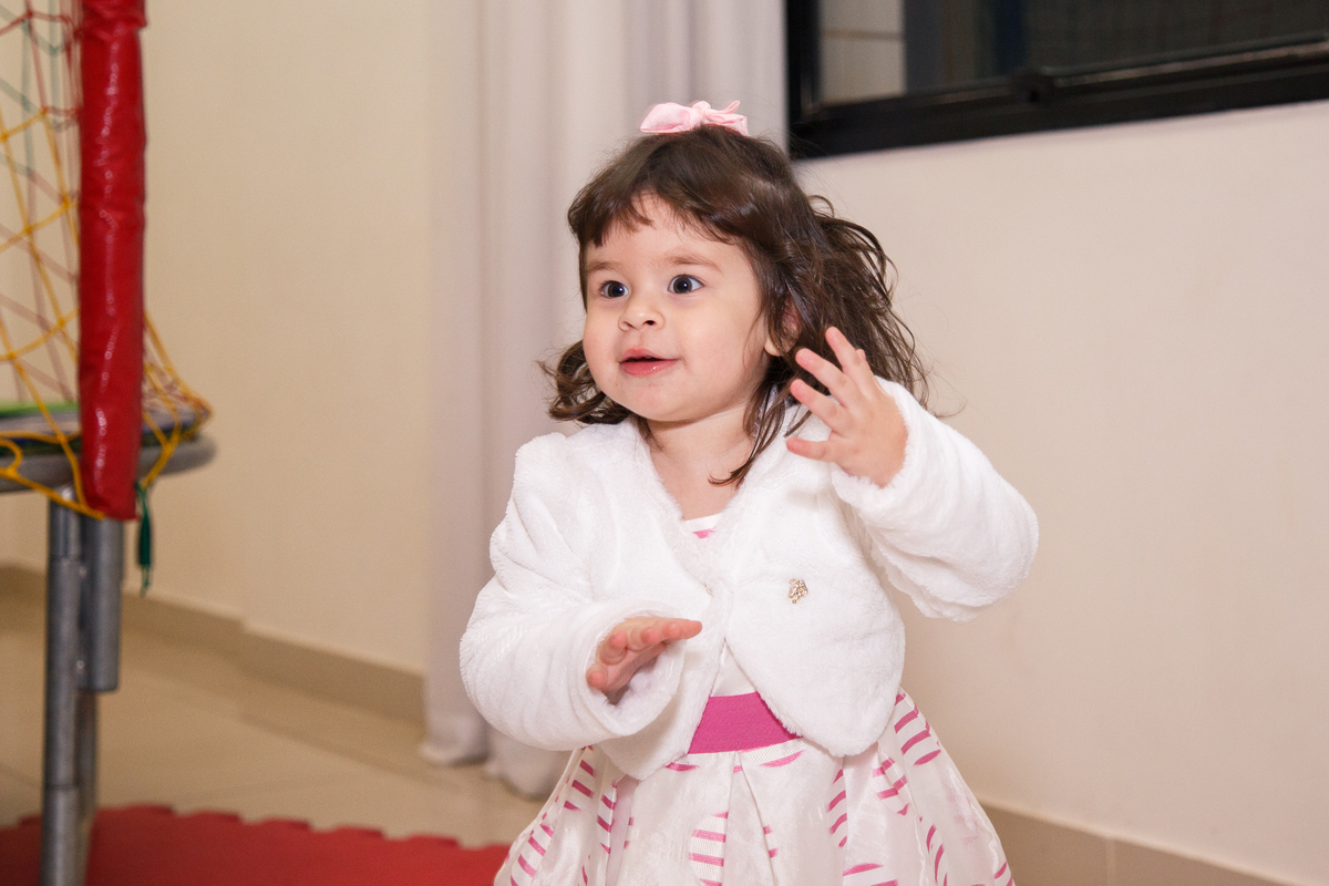 Menina de 2 anos dançando no meio da foto, usando um vestido branco com detalhes rosa e um casaquinho branco.