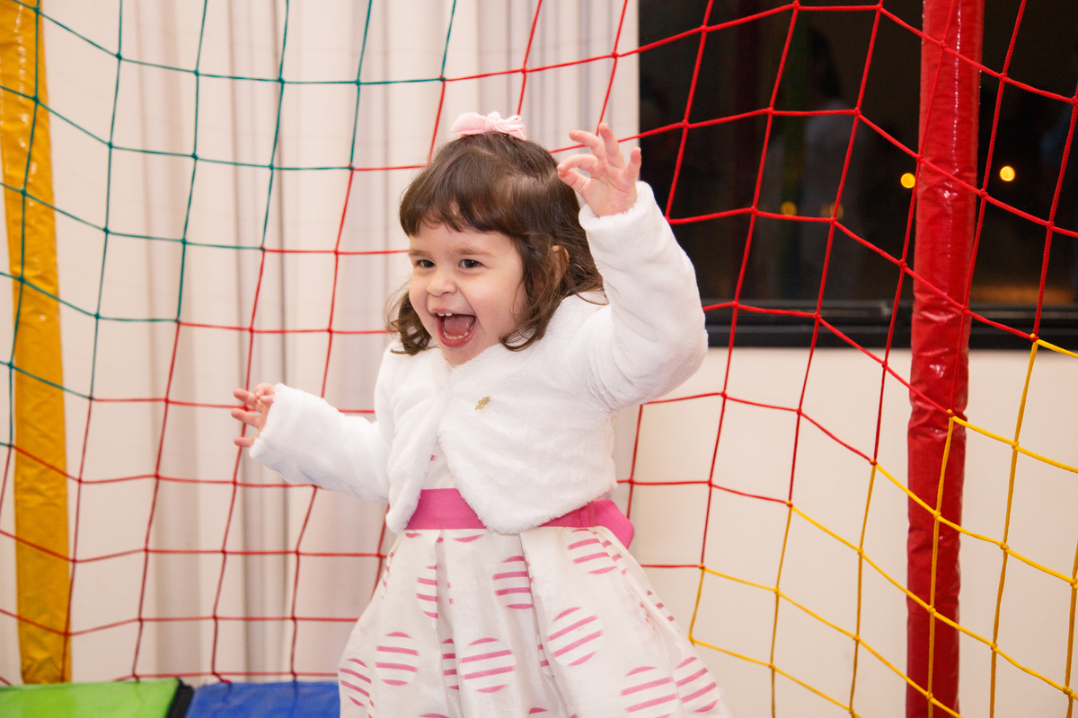 Menina de 2 anos pulando no pula pula e dando um grito de alegria, usando um vestido branco com detalhes rosa e um casaquinho branco.