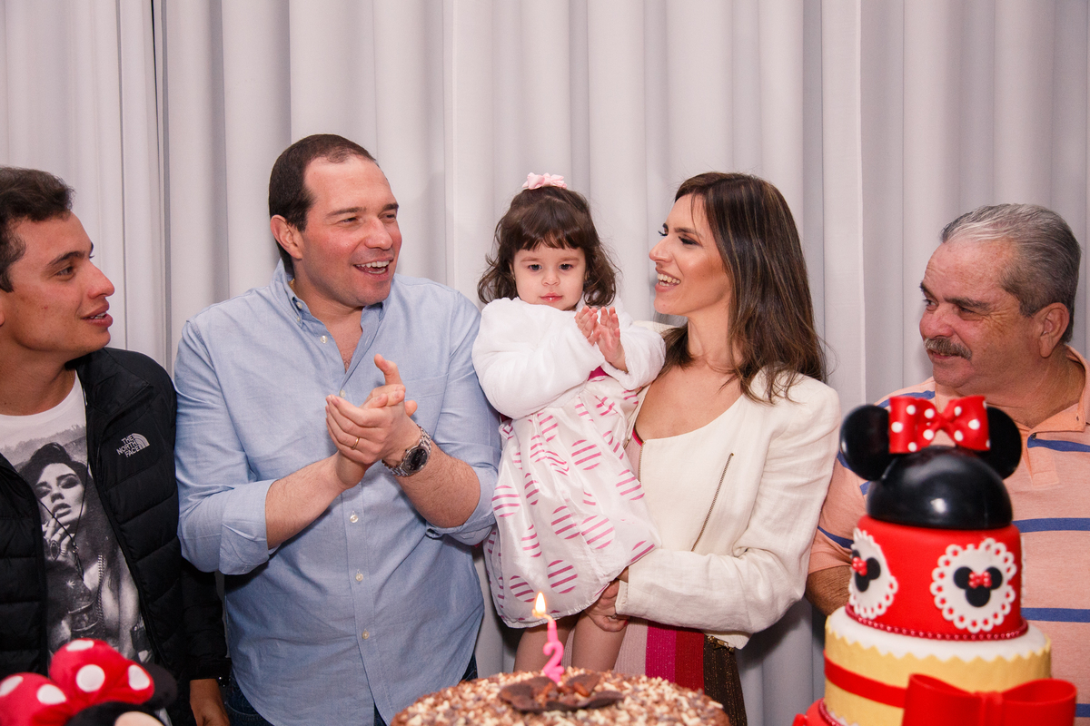 Mesa do bolo na hora do parabéns para menina de 2 anos. Ela no colo da mãe, ao lado o pai, avô e tio, todos atrás da mesa do bolo.
