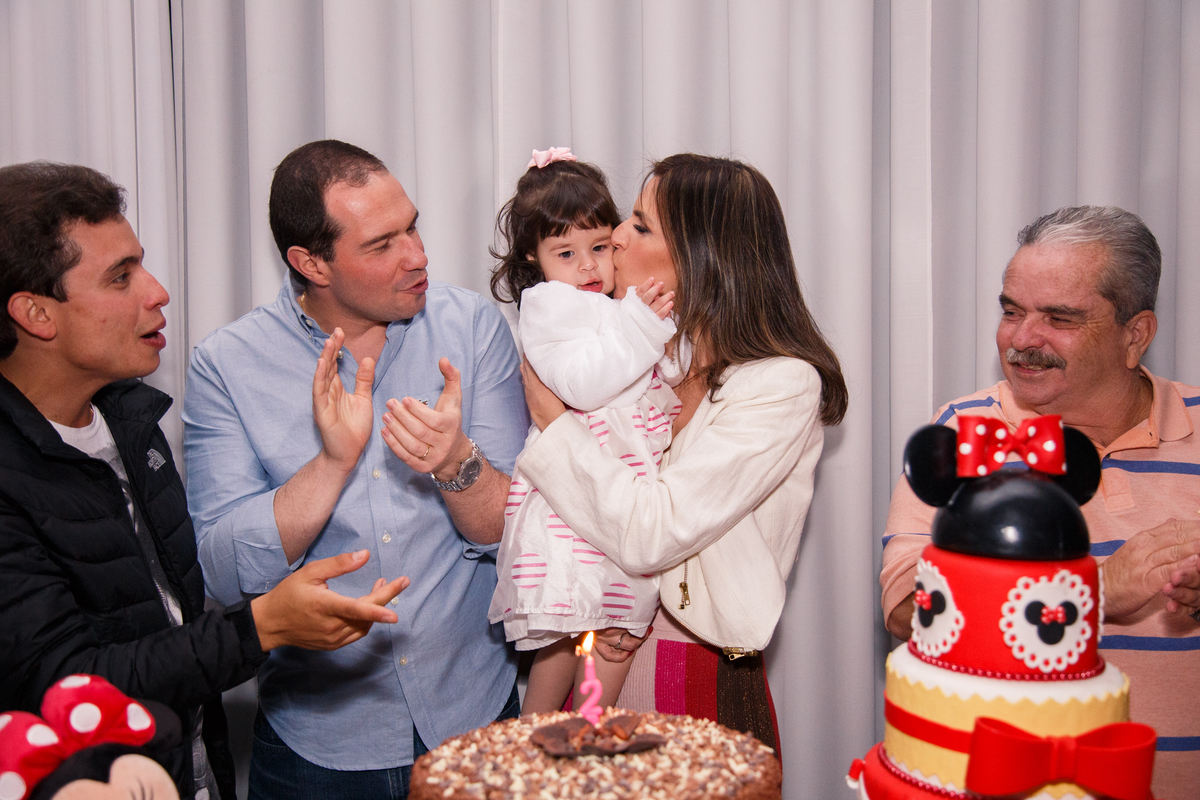 Mesa do bolo na hora do parabéns para menina de 2 anos. Ela no colo da mãe, ganhando um beijo na bochecha, ao lado o pai, avô e tio, todos atrás da mesa do bolo.