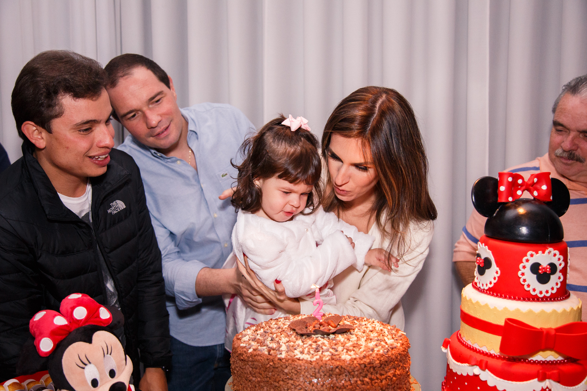 Mesa do bolo na hora do parabéns para menina de 2 anos. Ela sopra a velinha no colo da mãe, ao lado o pai, avô e tio, todos atrás da mesa do bolo.