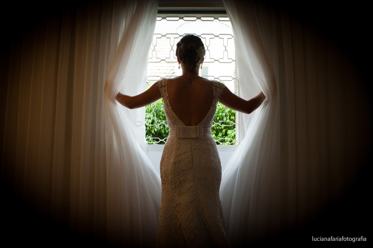 decote, costas de fora, vestido, coque baixo, cabelo de noiva, maquiagem, cabeleireiros, arrumando a noiva, vestido de renda, renda, flor, flor no cabelo, tatuagem, tatuagem nas costas, janela, foto na janela, silhueta, laço no vestido