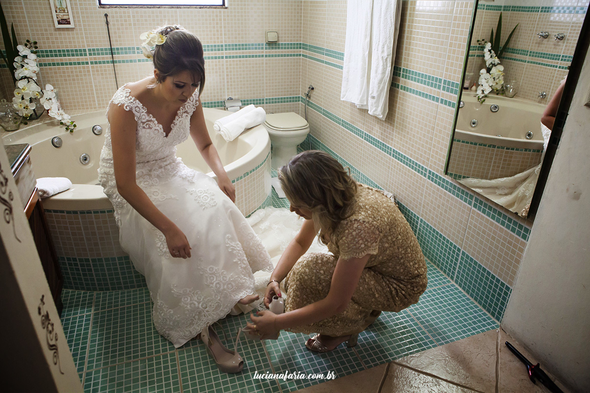dia da noiva em casa com a mãe abotoando o sapato da noiva em linda foto da fotógrafa de casamento luciana faria também fotografa de família