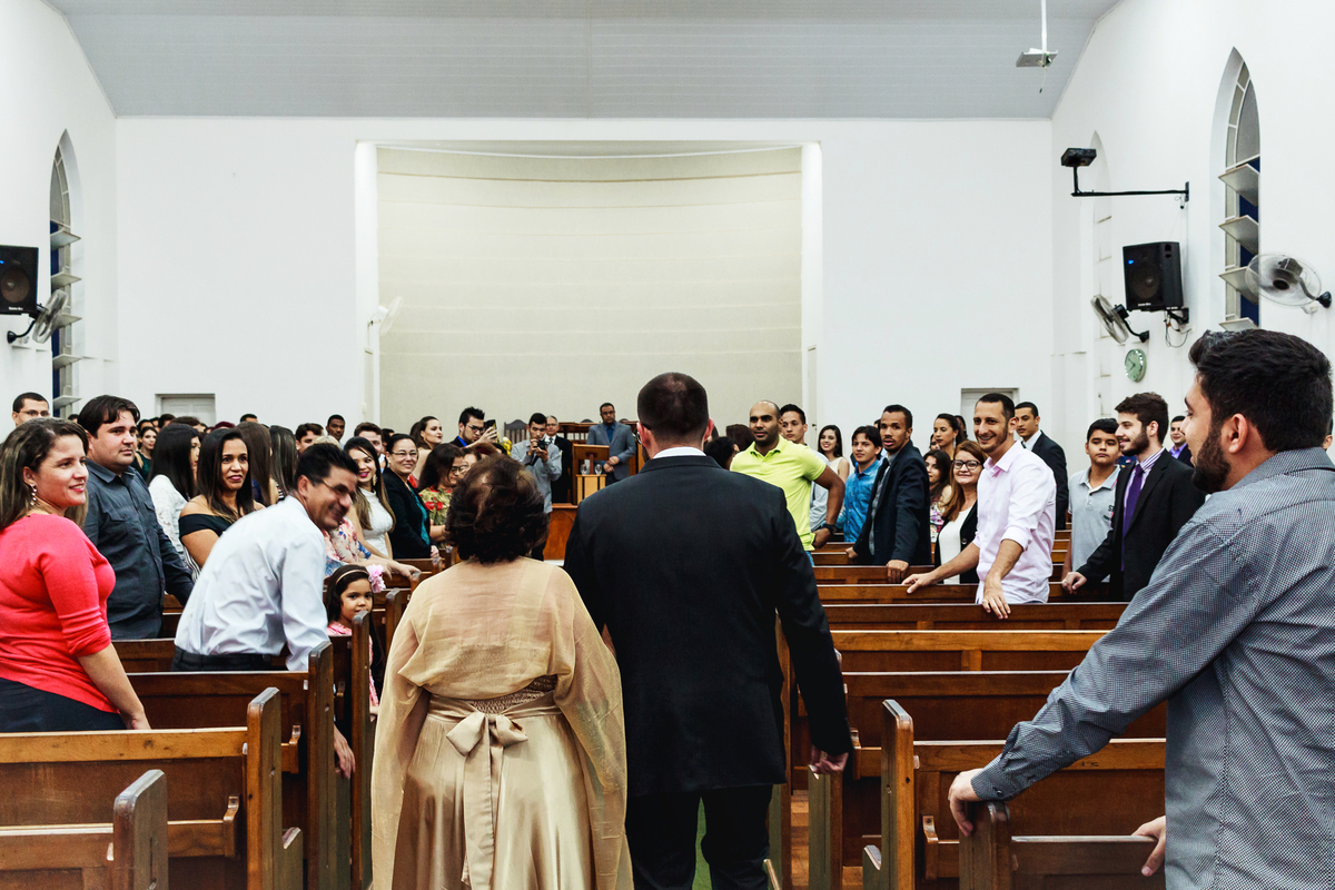 Noivo e mãe do noivo entrando na igreja branca com várias pessoas sorrindo.
