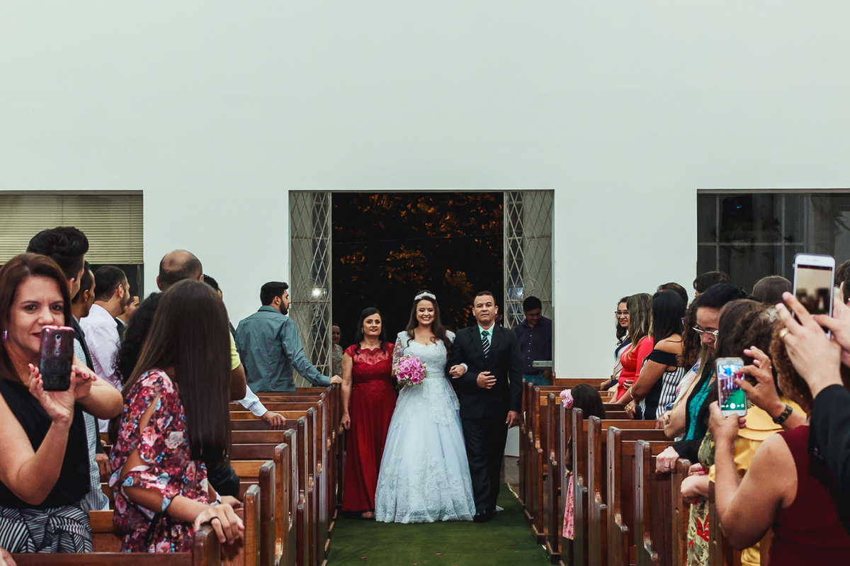 Noiva entrando com os pais pela porta da igreja, vestindo um vestido branco com um buquê de flores cor de rosa