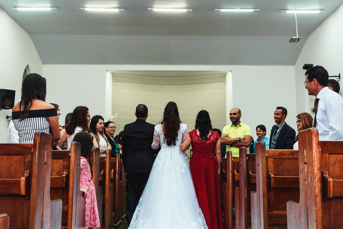 Noiva entrando com os pais pela porta da igreja, vestindo um vestido branco com um buquê de flores cor de rosa