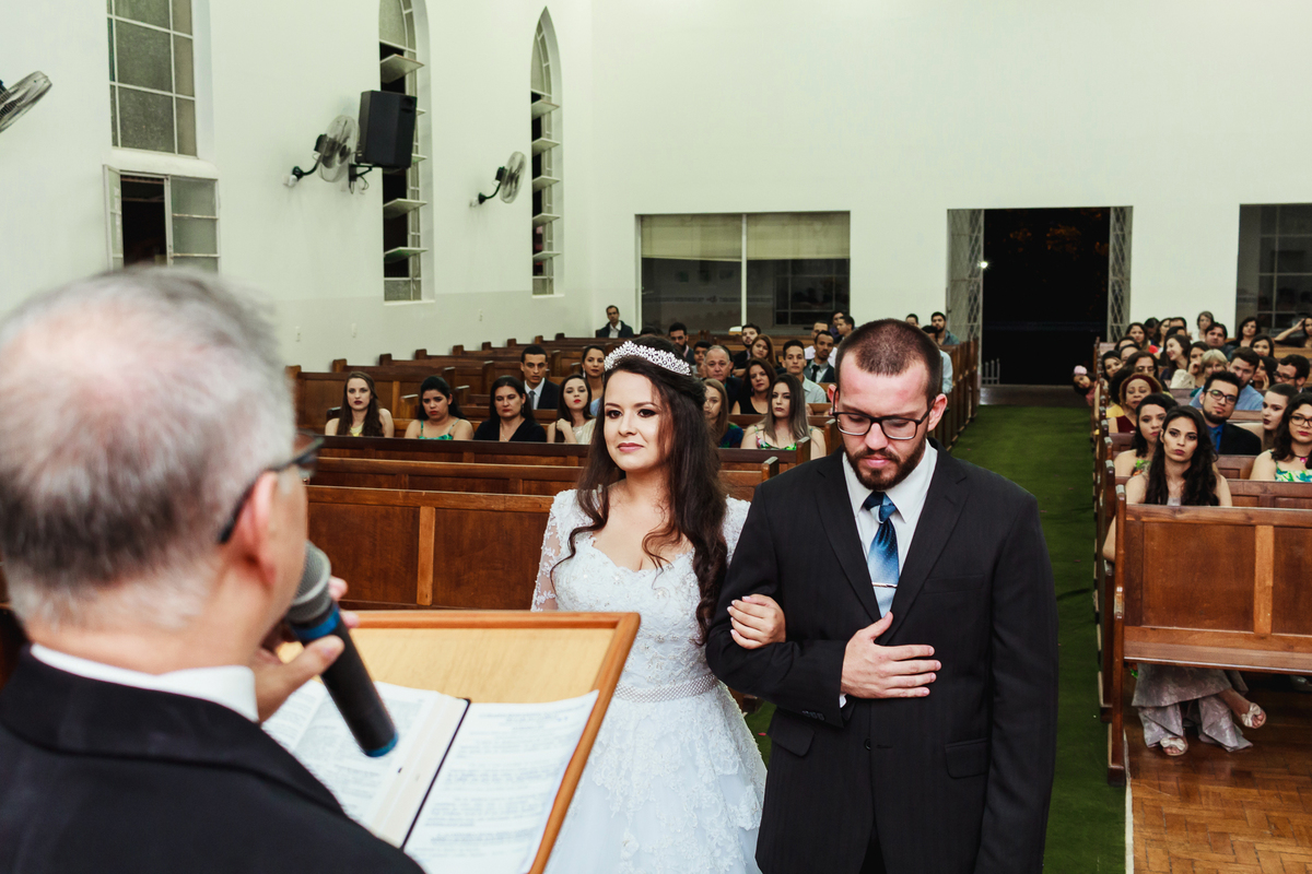 Noivo vestindo um vestido  segurando um buquê de flores cor de rosa ao lado do noivo que veste um lindo terno de casamento com um pastor falando ao microfone