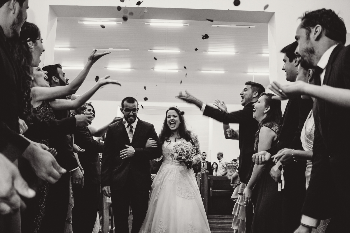 Noivos saindo da igreja com pétalas de rosas lançadas ao ar, sorrindo com o momento divertido e emocionante durante o casamento.