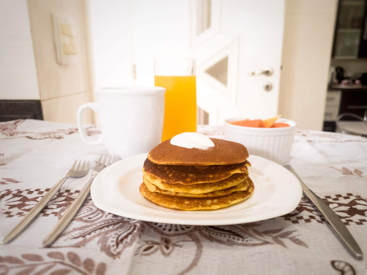 mesa com panquecas de fubá com requeijão no prato, caneca, talheres, ramequim com frutas e copo de suco de laranja