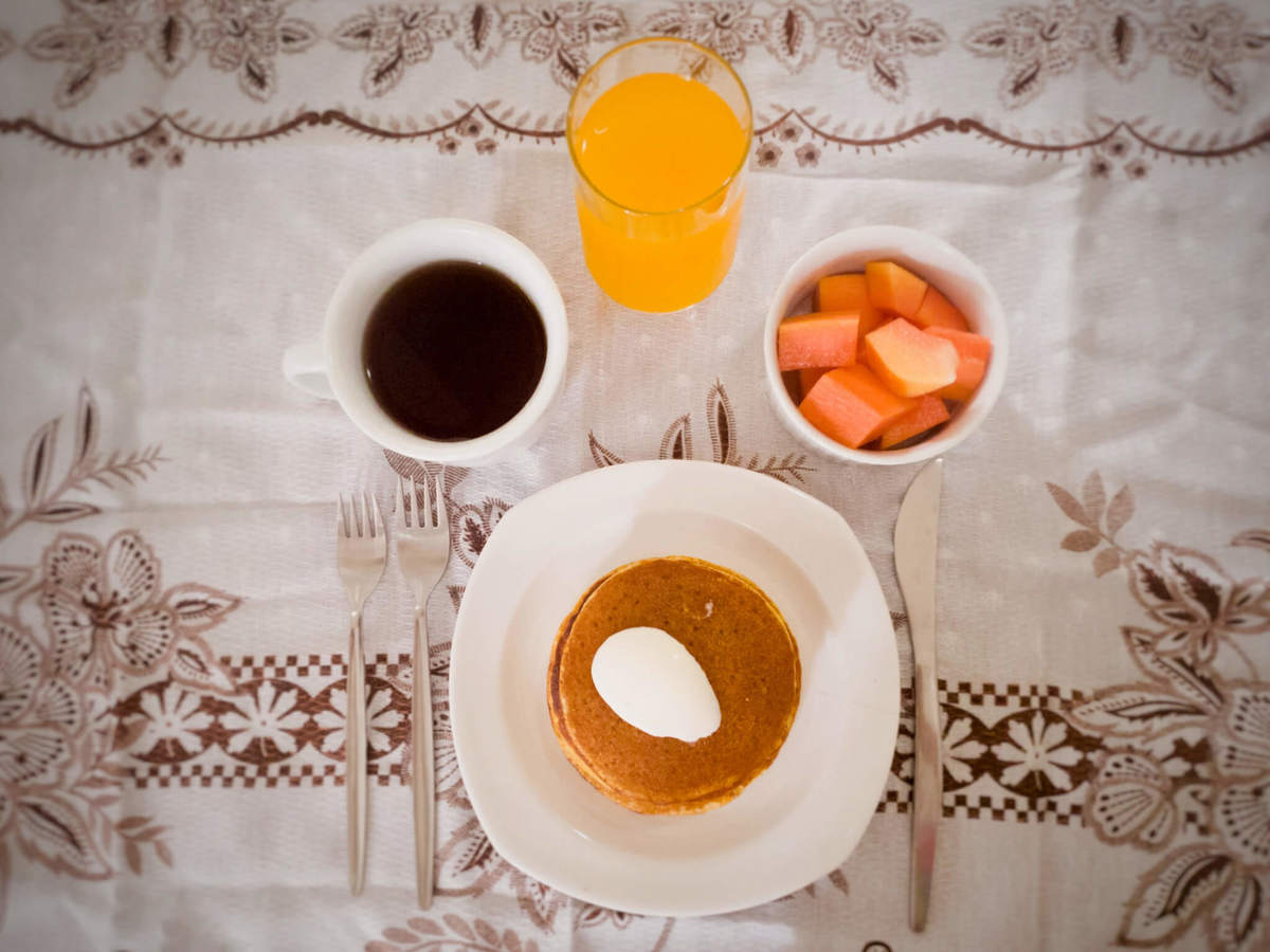 mesa com panquecas de fubá com requeijão no prato, caneca com chá, talheres, ramequim com frutas e copo de suco de laranja vista de cima